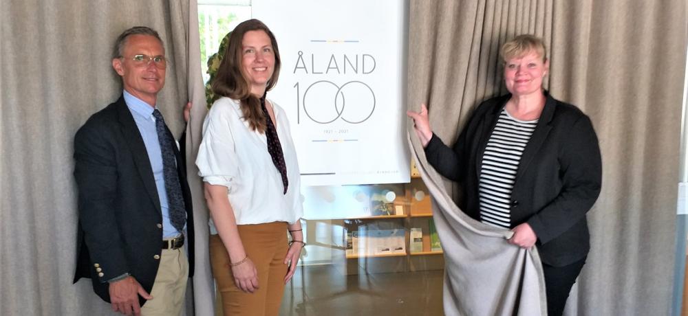 Stefan Aspberg projektledare för Åland 100, Annika Zetterqvist vinnare och lantrådet Katrin Sjögren vid bilden av logotypen.