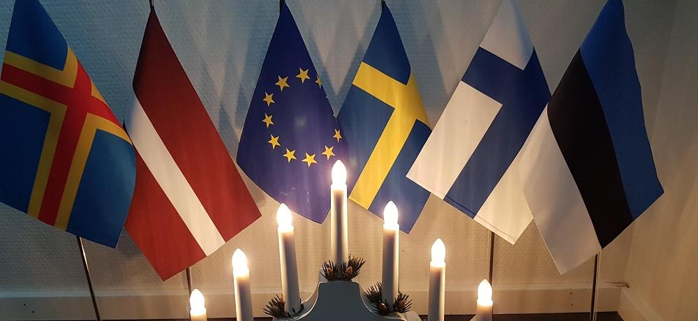 Ålands, Lettlands, EU:s, Sveriges, Finlands och Estlands flaggor som bordsflaggor bakom en tänd adventsljusstake.