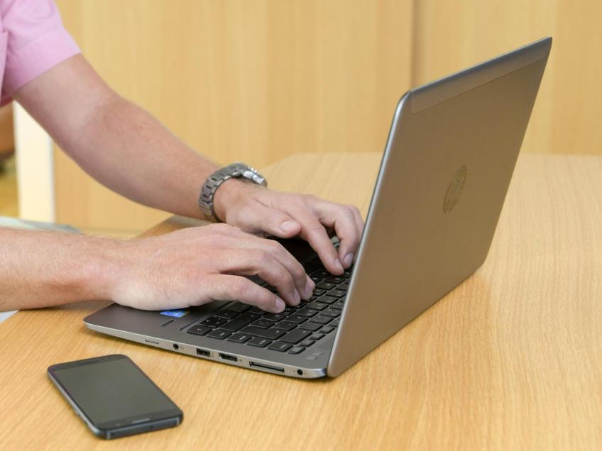 Händer som skriver på en bärbar dator, bredvid på bordet ligger en mobiltelefon.